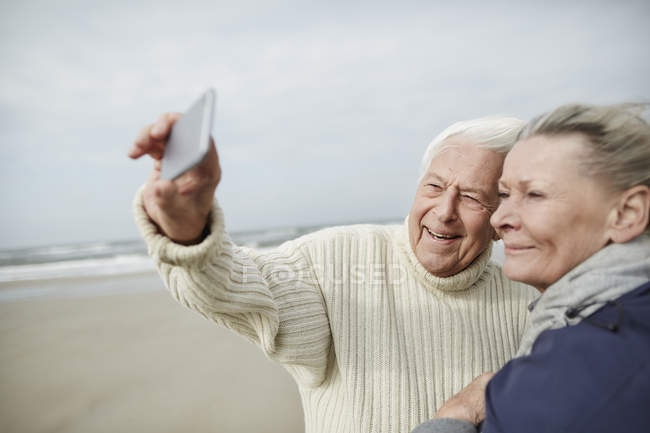 Coppia anziana scattare selfie con il cellulare sulla spiaggia invernale ventosa — Foto stock