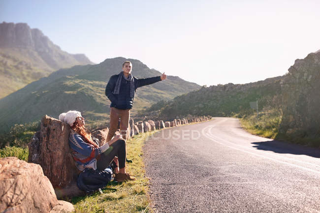 Young couple hitchhiking at sunny, remote roadside — Stock Photo