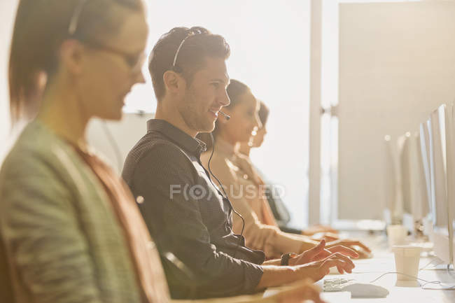 Telemarketers wearing headsets talking on telephone and working at computers in a row in sunny office — Stock Photo