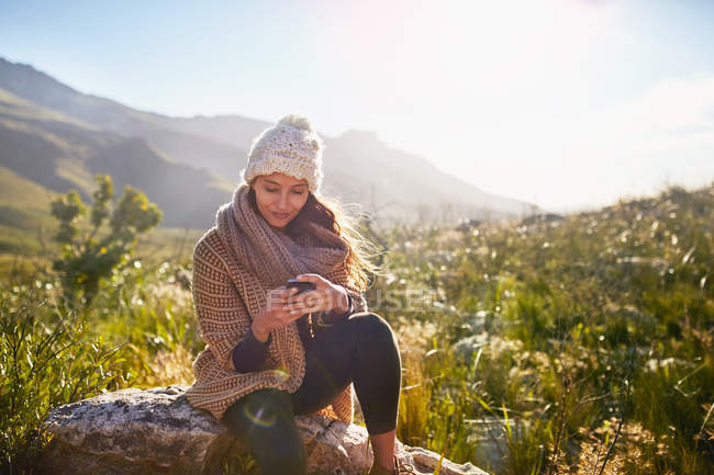 Молода жінка смс з мобільним телефоном на скелі в сонячному, віддаленому полі — стокове фото