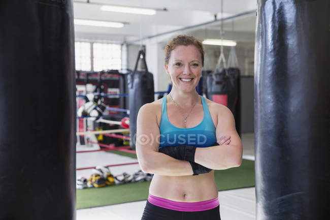 Улыбающаяся, уверенная в себе женщина-боксер, стоящая перед боксерскими грушами в спортзале — стоковое фото