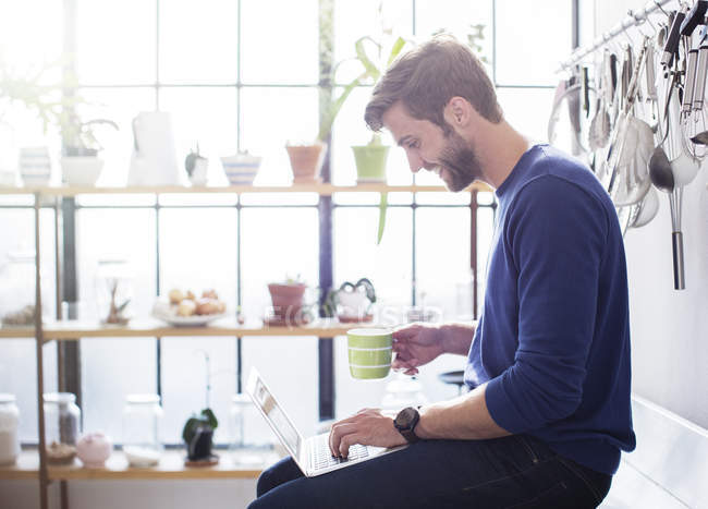 Young man sitting on kitchen counter with mug and laptop — Stock Photo