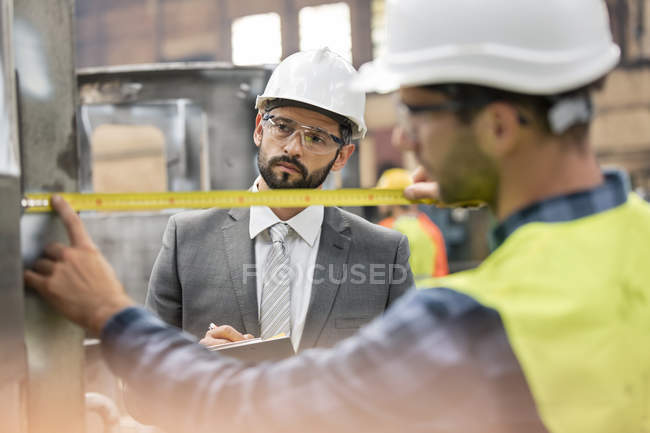 Manager watching steel worker with tape measure in factory — Stock Photo