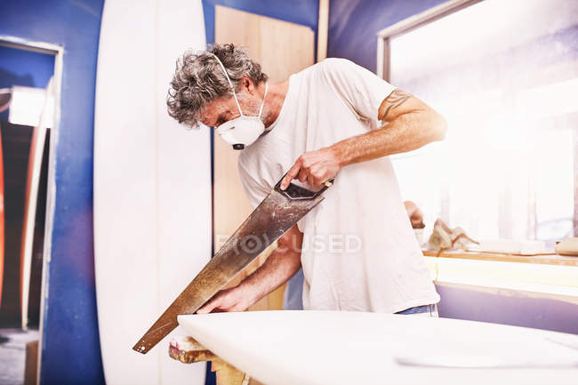 Male surfboard designer wearing protective mask and using saw in workshop — Stock Photo