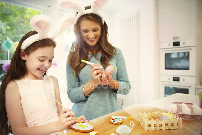 Мать и дочь в костюме кролика уши окрашивая пасхальные яйца и печенье — стоковое фото