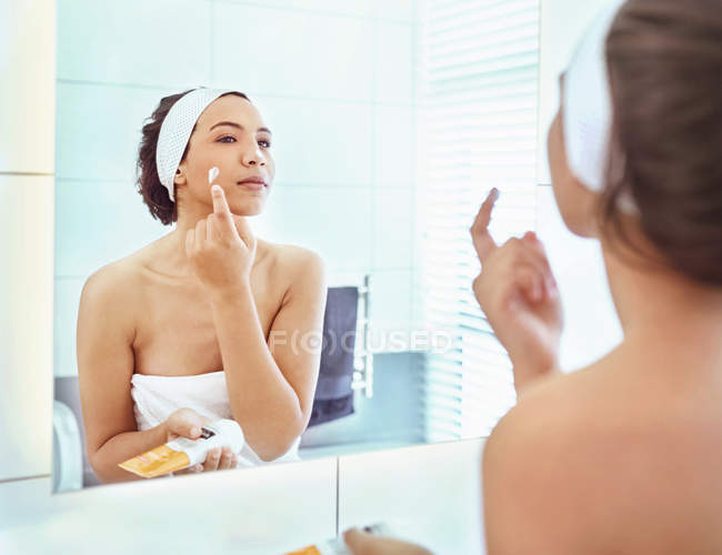 Woman applying moisturizer to cheek at bathroom mirror — Stock Photo