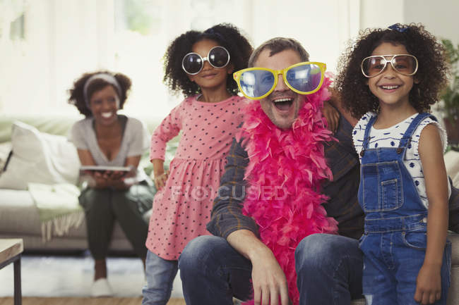 Retrato juguetón multi-étnico padre e hijas jugando a disfrazarse con gafas de sol y boas de plumas - foto de stock