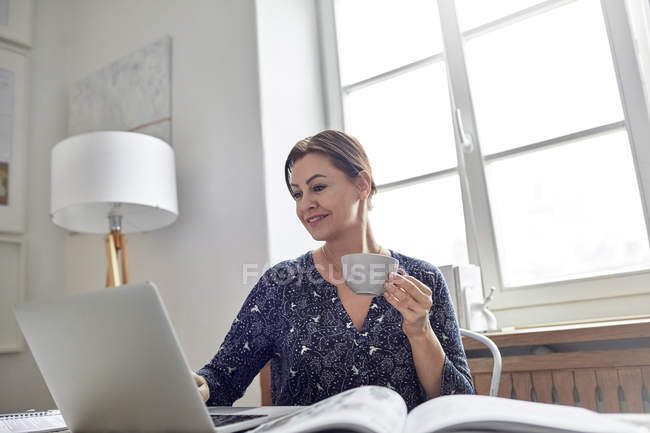 Femme d'affaires souriante buvant du café et travaillant à l'ordinateur portable — Photo de stock