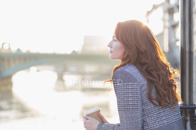 Femme d'affaires sereine aux cheveux roux buvant du café au bord de l'eau urbain — Photo de stock