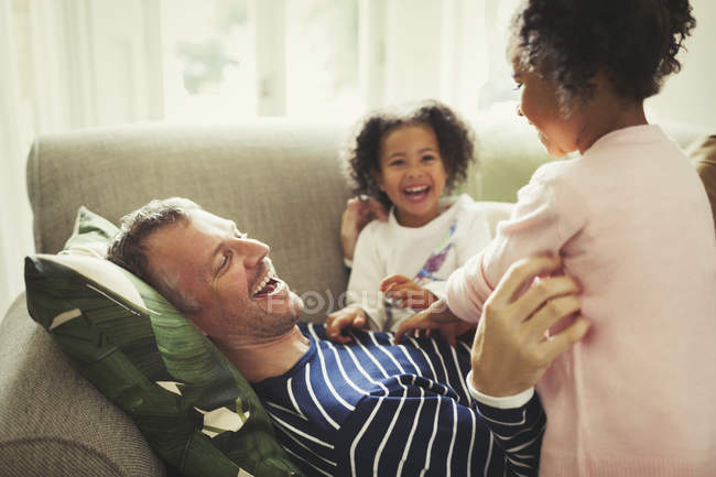 Juguetón multi-étnico padre e hijas cosquillas y riendo en el sofá - foto de stock