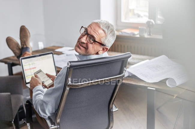 Portrait homme d'affaires souriant et confiant avec téléphone portable et ordinateur portable au bureau — Photo de stock