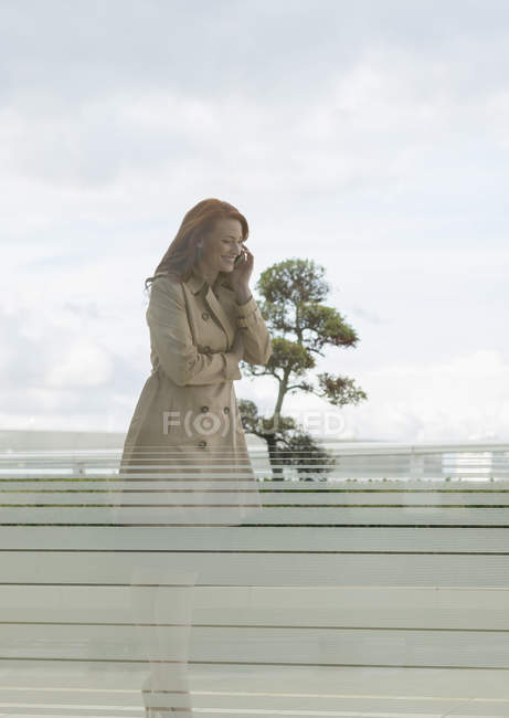 Улыбающаяся деловая женщина с рыжими волосами разговаривает по мобильному телефону на балконе — стоковое фото