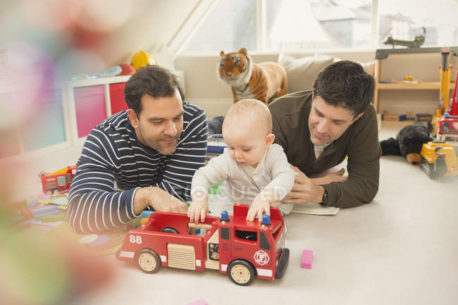 Male gay parents and baby son playing with fire engine toy in playroom — Stock Photo
