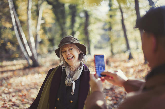 Verspielte Seniorin wird im Herbstpark von Ehemann mit Kameratelefon fotografiert — Stockfoto