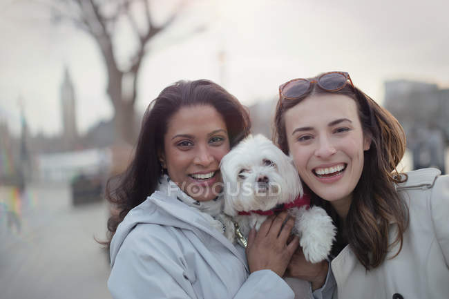Retrato sonriente pareja lesbiana con perro blanco en parque urbano - foto de stock
