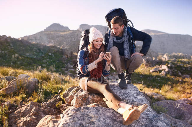 Young couple with backpacks hiking, resting on rock using smart phone — Stock Photo