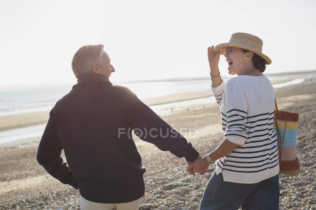 Laughing mature couple holding hands and walking on sunny beach — Stock Photo