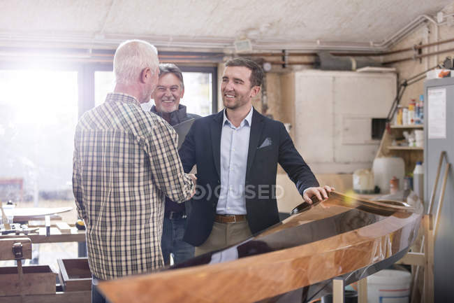 Carpinteros machos apretón de manos con cliente satisfecho junto a madera kayak en taller - foto de stock
