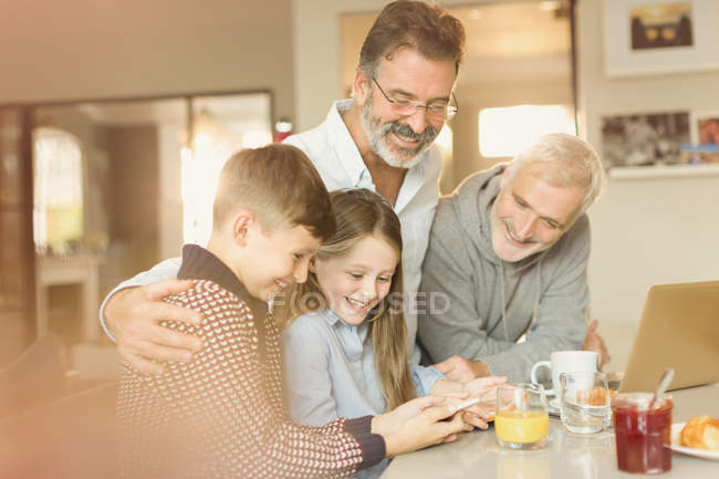 Hombres padres gay y niños usando el teléfono celular en el mostrador de la cocina - foto de stock
