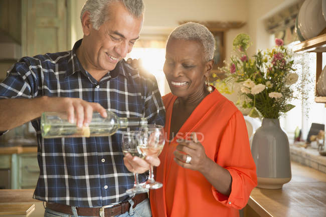 Sonriente pareja de ancianos vertiendo vino blanco en la cocina - foto de stock