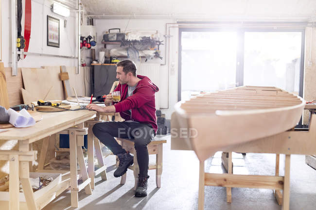 Мужчина плотник пьет чай и работает за ноутбуком на рабочем столе в мастерской — стоковое фото