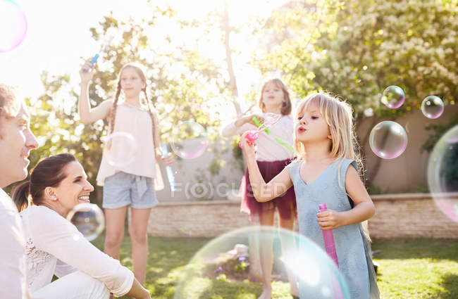 Pais assistindo filhas soprando bolhas no quintal ensolarado — Fotografia de Stock
