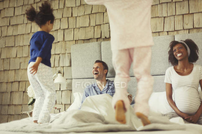 Multi-ethnic pregnant parents watching daughters jumping on bed — Stock Photo