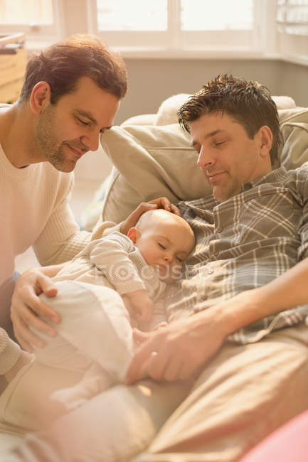 Male gay parents watching baby son sleeping on sofa — Stock Photo