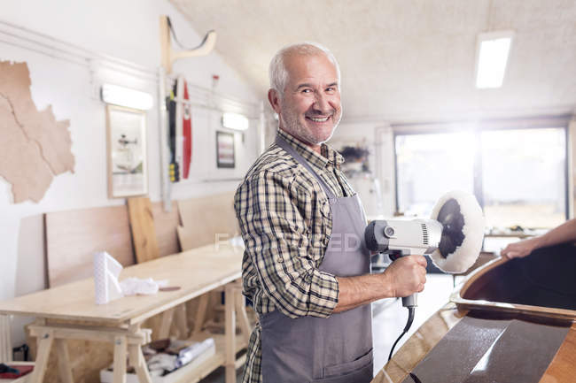 Retrato sorridente, confiante carpinteiro masculino sênior usando uma lixadeira tampão em barco de madeira na oficina — Fotografia de Stock