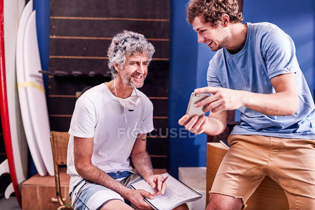 Male surfboard designers with cell phone brainstorming in workshop — Stock Photo
