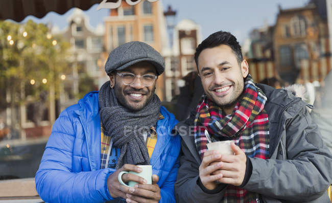Портрет улыбающихся молодых людей в теплой одежде, пьющих кофе в городском кафе на тротуаре — стоковое фото