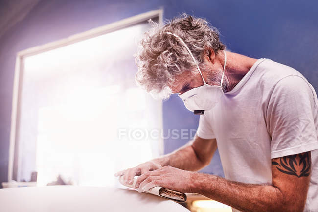 Hombre enfocado con máscara protectora lijando tabla de surf en taller - foto de stock