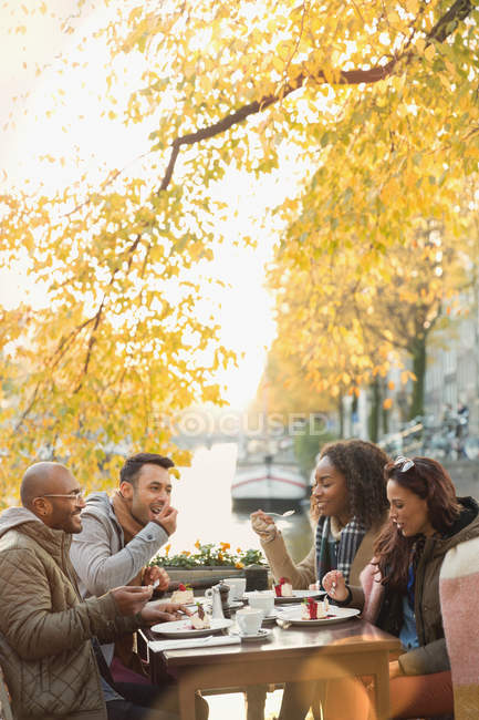 Amigos comiendo postre y tomando café en la acera de otoño café a lo largo del canal - foto de stock