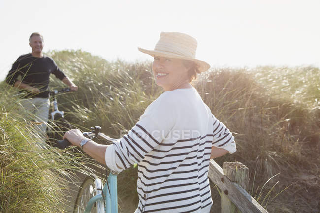 Porträt lächelnde ältere Frau zu Fuß Fahrrad am sonnigen Strand Grasweg — Stockfoto