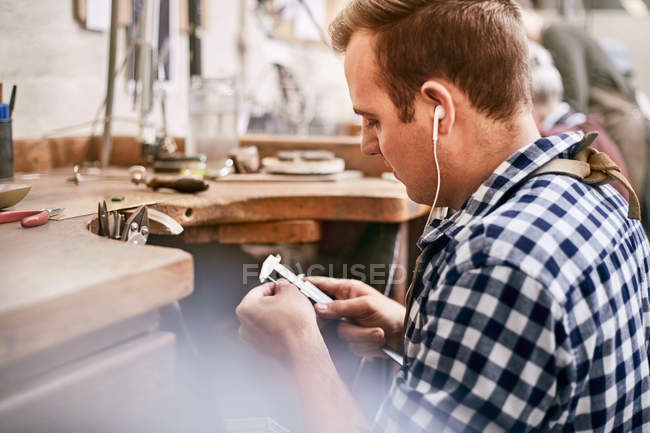 Male jeweler using calipers and listening to music with headphones in workshop — Stock Photo