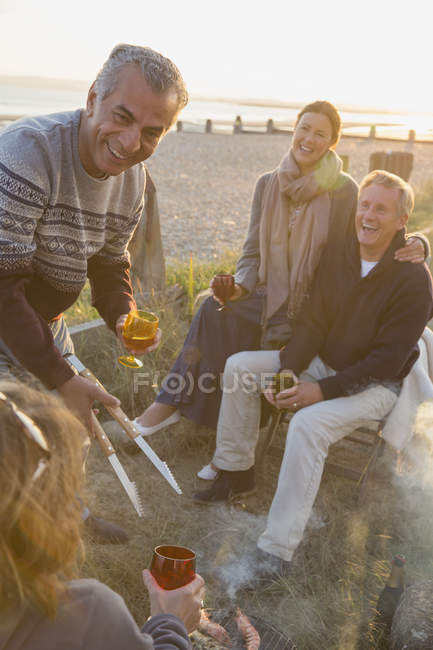Parejas maduras sonrientes tomando vino y haciendo barbacoas en la playa al atardecer - foto de stock