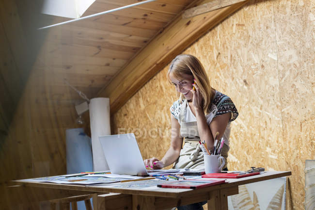 Smiling artist talking on cell phone and using laptop in studio — Stock Photo