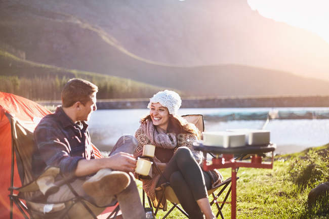 Sorrindo jovem casal bebendo café no acampamento ensolarado à beira do lago — Fotografia de Stock
