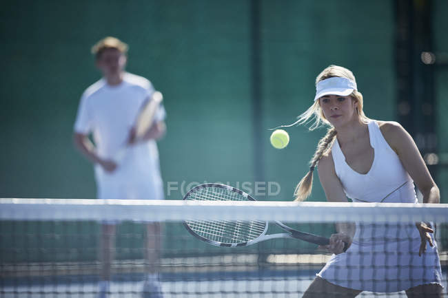 Determined young female tennis player playing tennis, hitting the ball at tennis net on sunny tennis court — Stock Photo