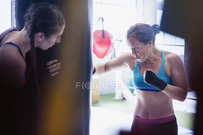 Boxeadores femeninos decididos y duros boxeando en el saco de boxeo en el gimnasio - foto de stock
