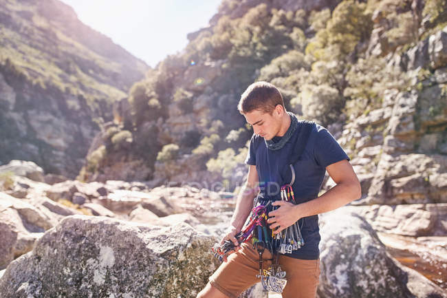 Hombre joven preparando mosquetones de escalada en roca y equipo entre rocas soleadas - foto de stock