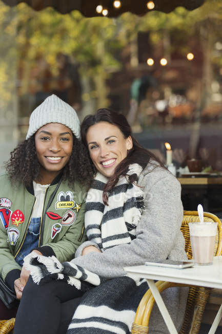 Портрет улыбающихся молодых женщин в теплой одежде в кафе на тротуаре — стоковое фото