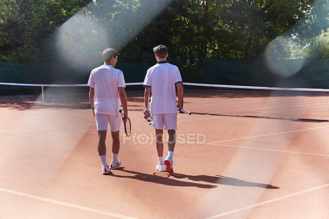 Jovens tenistas do sexo masculino caminhando com raquetes de tênis na quadra de tênis de barro ensolarado — Fotografia de Stock