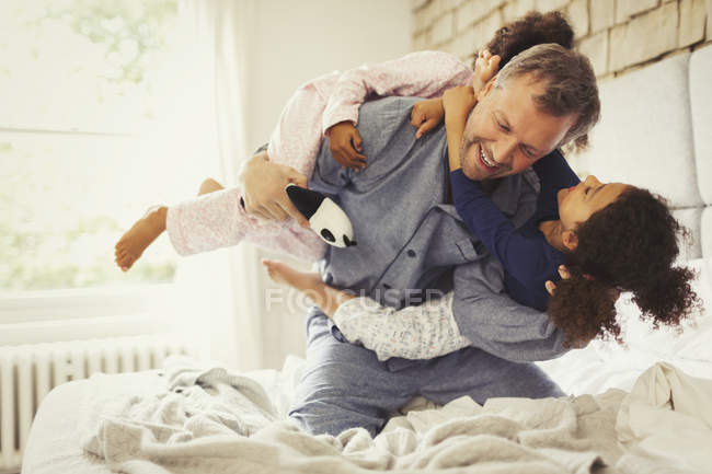 Multi-ethnic daughters playing and tackling father on bed — Stock Photo