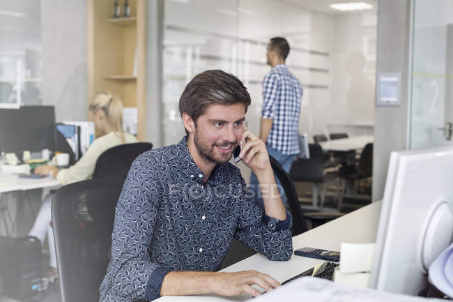 Бизнесмен разговаривает по мобильному телефону, работает за компьютером в офисе — стоковое фото