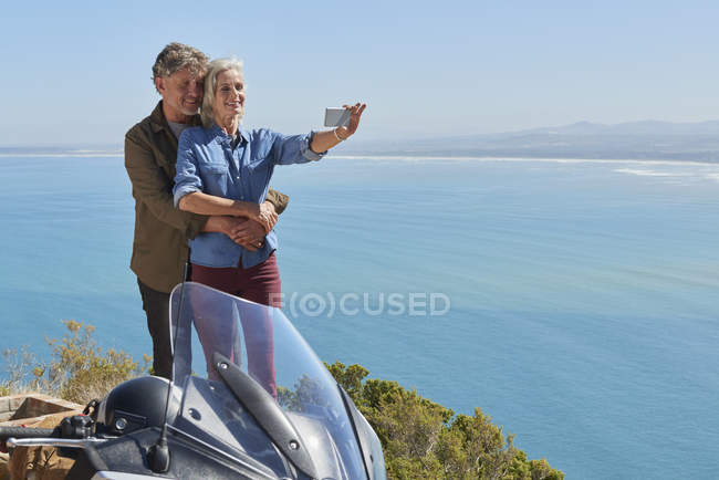 Casal sênior tirando selfie ao lado da motocicleta com vista para o mar ensolarado — Fotografia de Stock