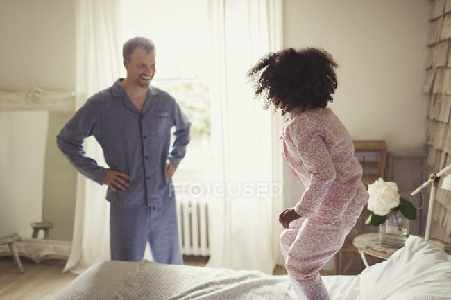 Багатоетнічний батько у піжамі дивиться, як дочка стрибає на ліжко — стокове фото