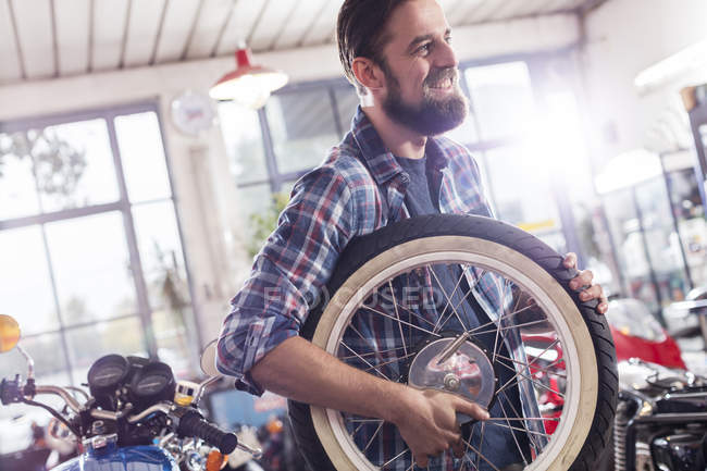 Smiling motorcycle mechanic carrying wheel in shop — Stock Photo