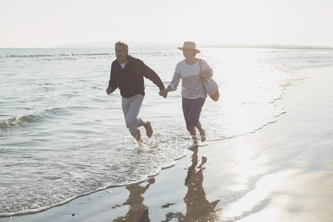 Brincalhão casal maduro de mãos dadas e correndo em ensolarado oceano surf — Fotografia de Stock