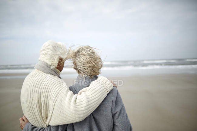 Pensive senior couple hugging and looking at ocean view on windy winter beach — Stock Photo